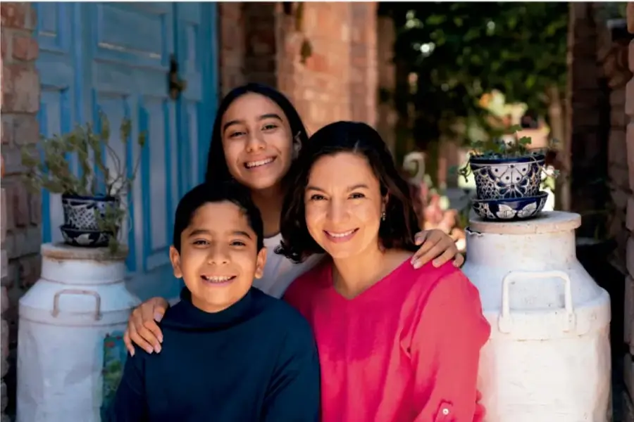 Fotografía de una madre con una niña y un niño sonriendo, junto a un muro de ladrillo, una puerta azul y dos globos de leche blancos con macetas decoradas encima.