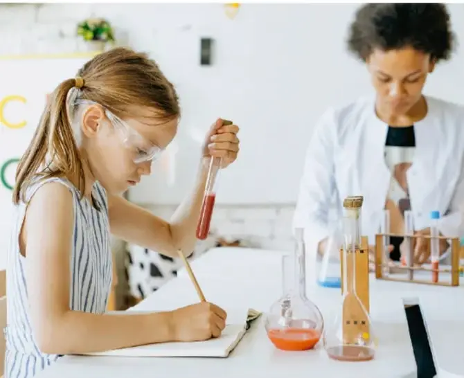 Fotografía de una estudiante con gafas de seguridad trabajando con matraces y tubos de ensayo en un laboratorio escolar, mientras otra persona observa al fondo.