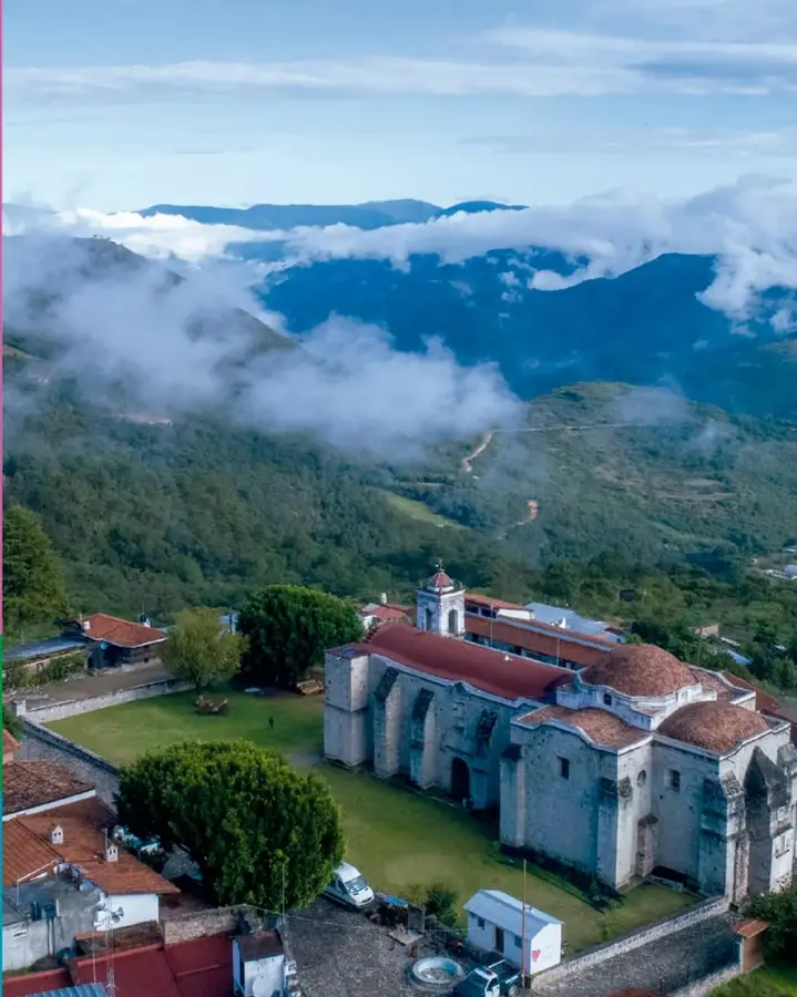 Fotografía aérea de un pueblo con un templo de cúpulas, pradera, casas con techo de teja y montañas cubiertas de nubes al fondo.