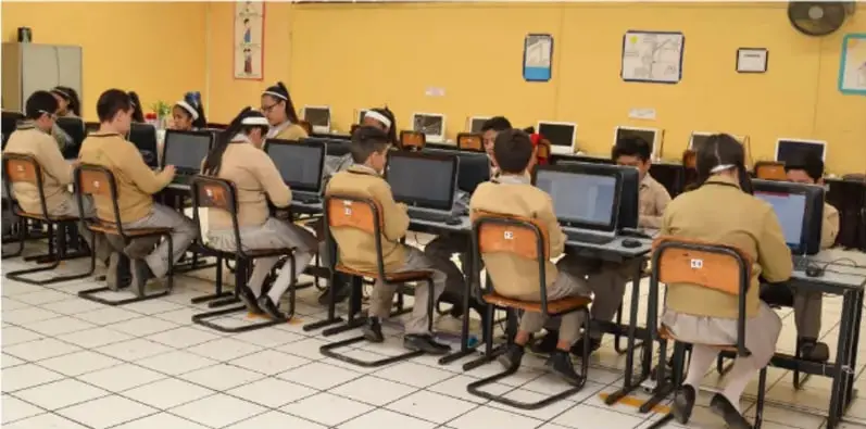Fotografía de estudiantes de secundaria en uniforme trabajando en una sala de cómputo frente a computadoras, realizando actividades del proyecto de modelación.