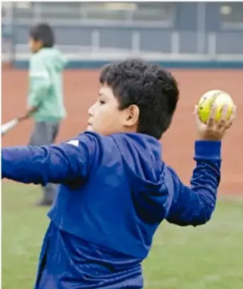 Niño con playera azul lanzando una pelota amarilla en una cancha, con otro niño al fondo.