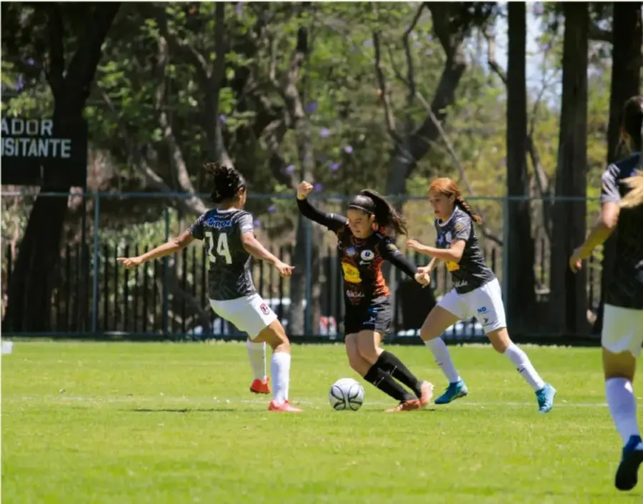 Fotografía de un partido de fútbol femenil en una cancha de pasto, con árboles al fondo y un letrero parcialmente visible que dice 'ADOR ISITANTE'; varias jugadoras disputan el balón.