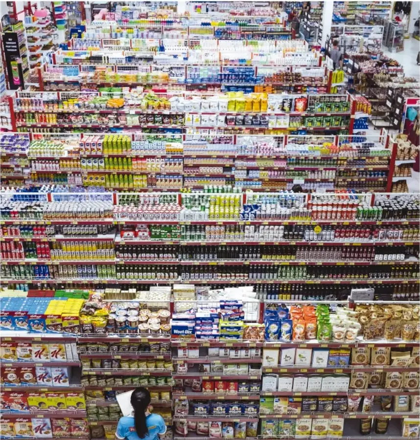 Fotografía de un supermercado con estantes llenos de productos variados y una persona de espaldas sosteniendo una lista.