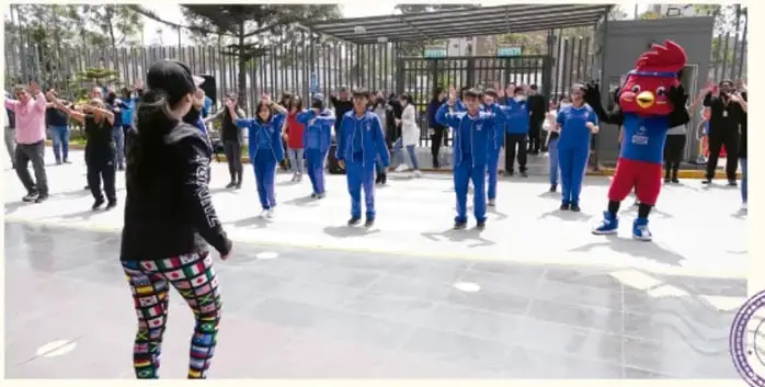 Fotografía de estudiantes en uniforme deportivo realizando una actividad física al aire libre con un docente y una mascota.