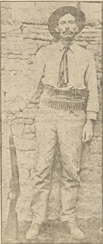 Fotografía en blanco y negro de Pascual Orozco de cuerpo entero, con sombrero de ala ancha, camisa clara, corbata, canana cruzada al pecho y carabina apoyada en el suelo.