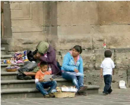 Fotografía de un grupo de personas, entre ellas niñas y niños, sentados en los escalones de un inmueble antiguo; parecen en situación de venta callejera o espera, ilustrando la desigualdad y la vulneración de derechos en el espacio público.