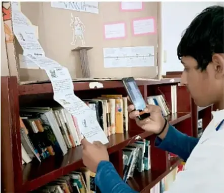 Estudiante tomando notas con teléfono junto a un estante de libros y un mural con recortes (crédito: Luis Eduardo García Medel, Tlaxcala).