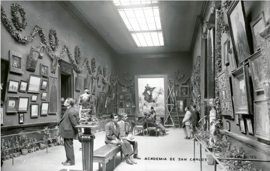 Fotografía en blanco y negro de una sala de museo con cuadros en las paredes, visitantes y una escalera; al pie se lee 'ACADEMIA DE SAN CARLOS'.