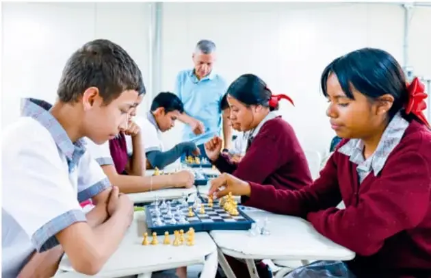 Fotografía de estudiantes de secundaria jugando ajedrez en mesas, con un docente observando al fondo; ilustra el trabajo colaborativo.