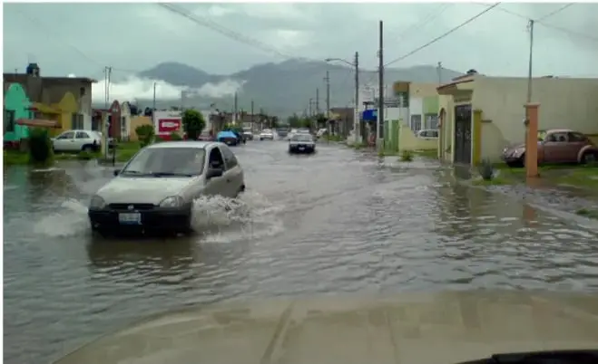 Fotografía de una calle inundada por agua acumulada; un automóvil avanza levantando salpicaduras. Al fondo se observan viviendas, postes de luz y montañas, lo que ilustra problemas de agua y drenaje en una comunidad.