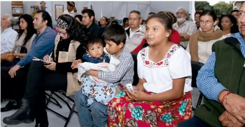 Fotografía de una reunión comunitaria: personas adultas, jóvenes y niños de distintas edades y culturas, algunas con vestimenta tradicional, sentadas escuchando una actividad.