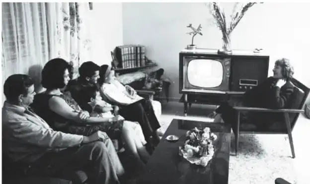 Fotografía en blanco y negro de una familia reunida en una sala viendo un televisor antiguo de tubo; se observan muebles de la época, un florero y cortinas, lo que ilustra la vida cotidiana del pasado.