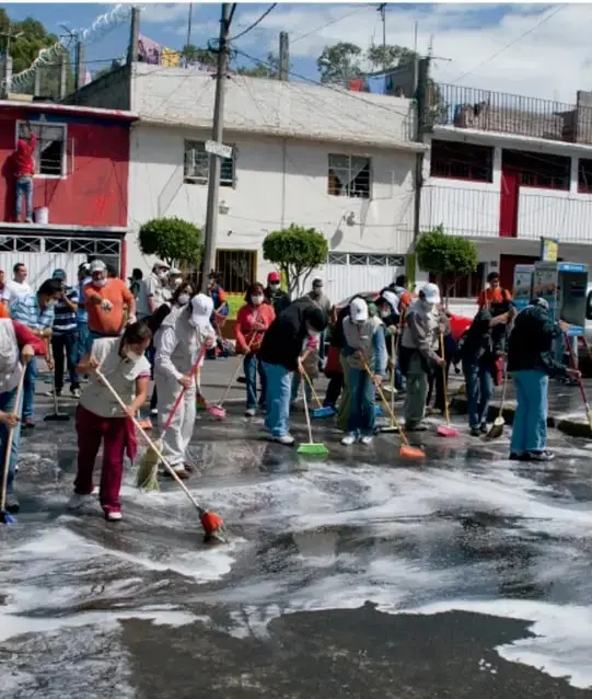 Fotografía de una comunidad realizando una jornada de limpieza en la calle, con personas usando escobas y cubetas sobre suelo enjabonado, en un entorno urbano de viviendas de dos niveles.