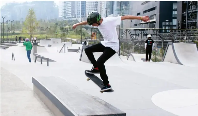 Fotografía de una persona con casco practicando skateboarding sobre un muro bajo en un skatepark urbano; al fondo hay rampas, otras personas observando y edificios de la ciudad.