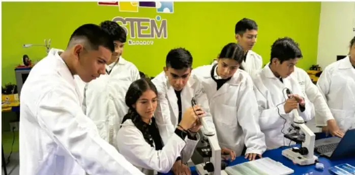 Fotografía de estudiantes con batas blancas (bata de laboratorio) en un aula con letrero STEM, trabajando con microscopios sobre una mesa azul, en el marco de la actividad del proyecto.