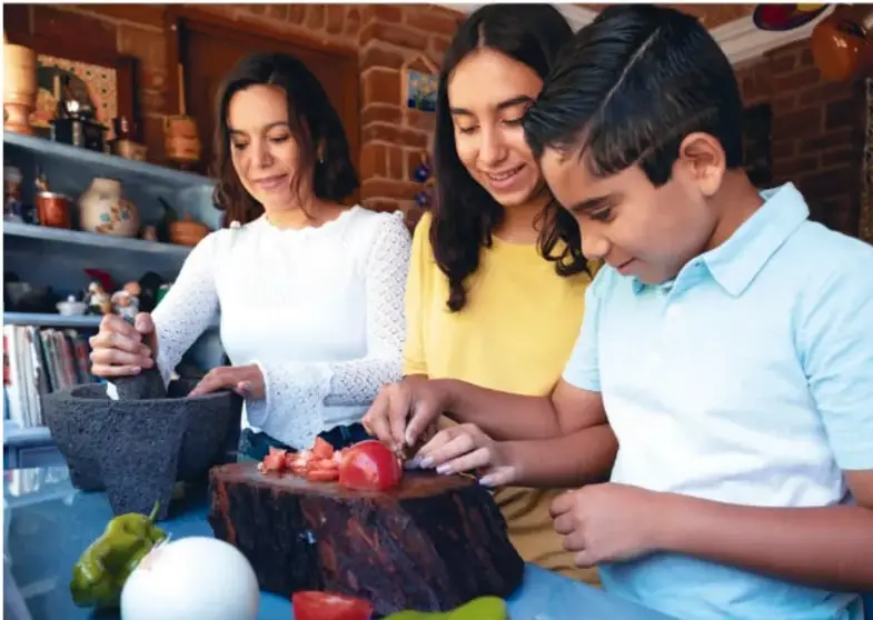 Fotografía de una madre y dos niños (una niña y un niño) preparando alimentos en una cocina: la madre usa un molcajete mientras los niños cortan un jitomate sobre una tabla.