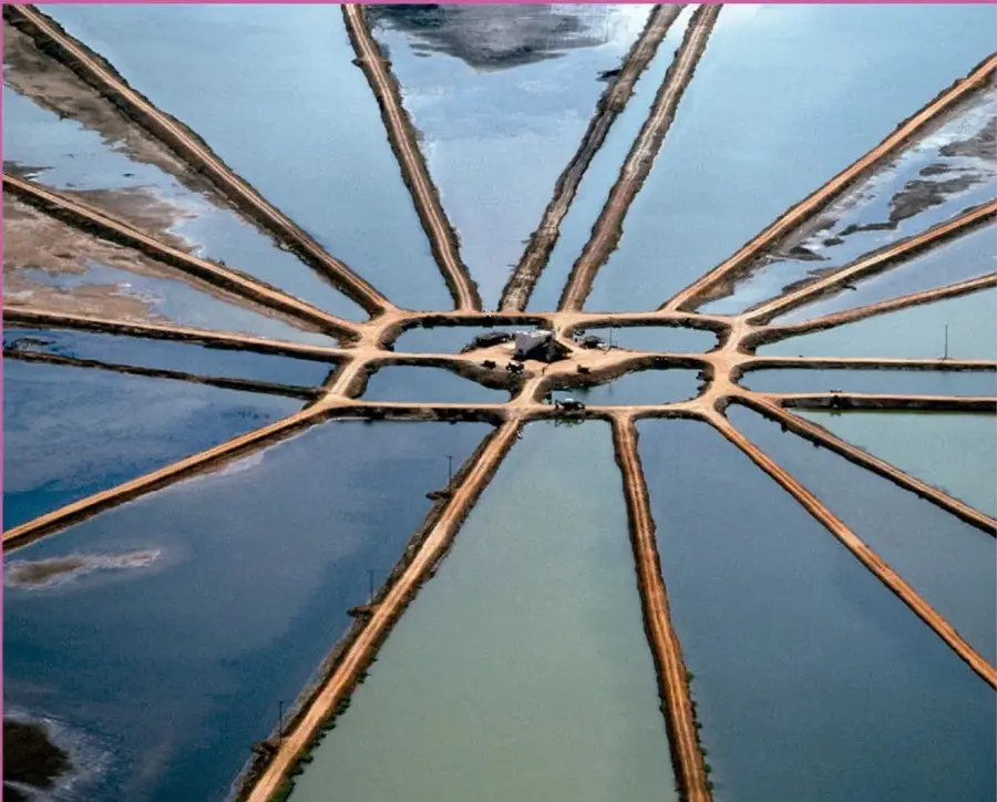 Fotografía aérea de salinas o estanques acuáticos divididos por terracerías que se distribuyen radialmente desde un punto central, con tonos azules y tierra.