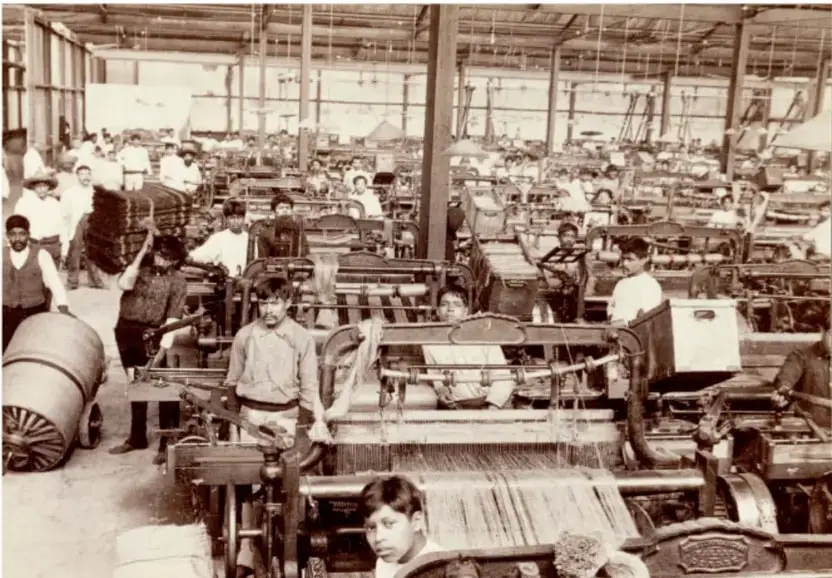 Fotografía en blanco y negro de una fábrica textil de principios del siglo XX. Se observan hileras de telares mecánicos, trabajadores adultos y niños operando las máquinas, y grandes carretes de hilo. Refleja la industrialización porfiriana y las condiciones laborales de la época.