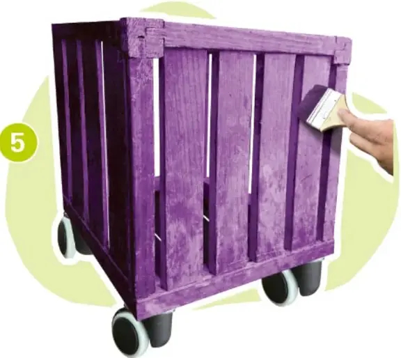 Fotografía de un huacal de madera pintado de morado con ruedas, sostenido por una mano que aplica pintura con brocha.