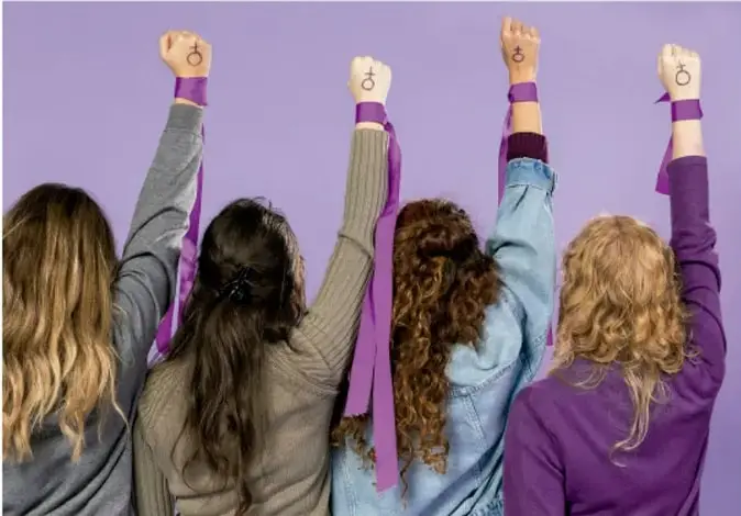 Fotografía de cuatro mujeres de espaldas con los puños levantados en señal de protesta, lucen pulseras y cintas moradas con el símbolo feminista (♀), sobre fondo lila.