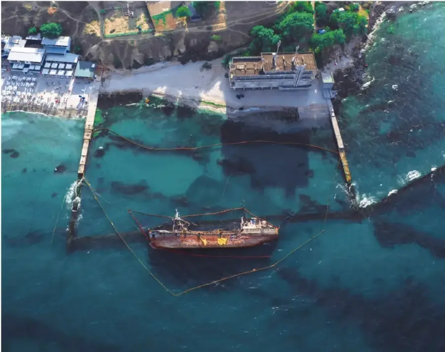 Fotografía aérea de una zona costera con un buque dentro de un área delimitada por barreras flotantes, rodeado de aguas turquesa; a un lado se observan instalaciones portuarias y una playa.