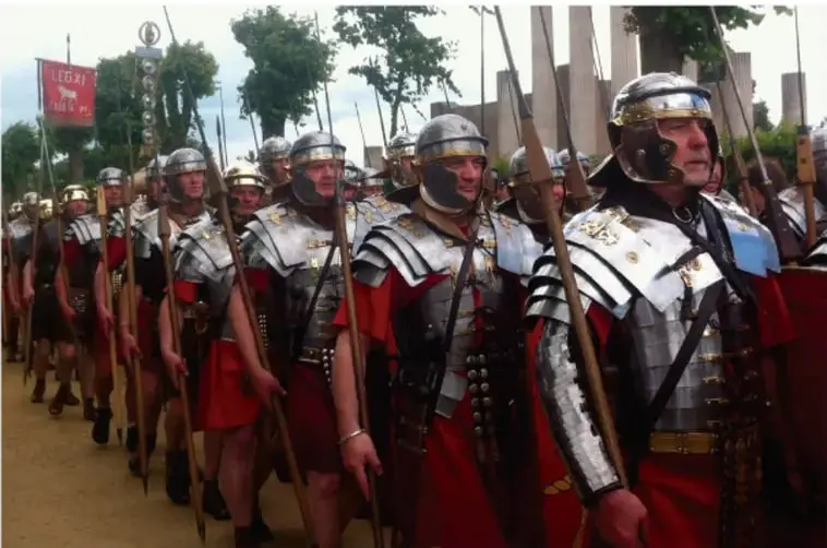 Fotografía de una recreación de soldados romanos (legionarios) marchando en formación, con armaduras, cascos, lanzas y estandartes.