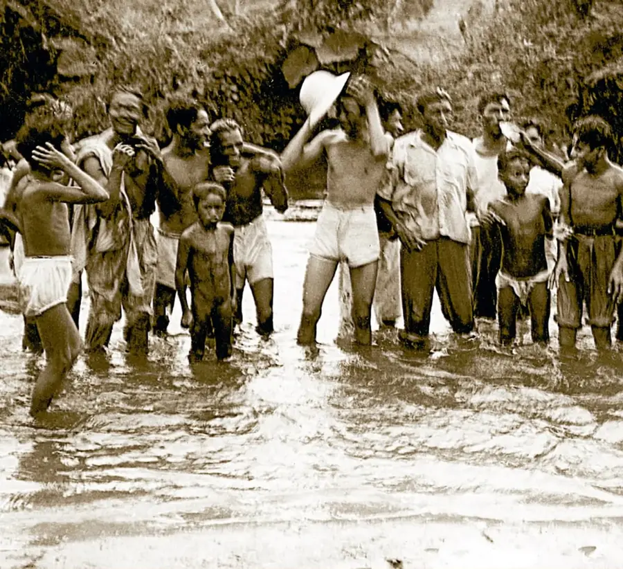 Fotografía sepia de época (ca. 1930) que muestra a un grupo de hombres, mujeres y niños de pie en un río o zona encharcada de la Costa Chica de Guerrero; varios llevan ropa ligera o semidesnudos, uno sostiene un sombrero en alto. Pie de foto: «Gente de la Costa Chica de Guerrero, ca. 1930. Archivo Gráfico de El Nacional, Fondo Temático, sobre: 01012-A (017). Acervo INEHRM».