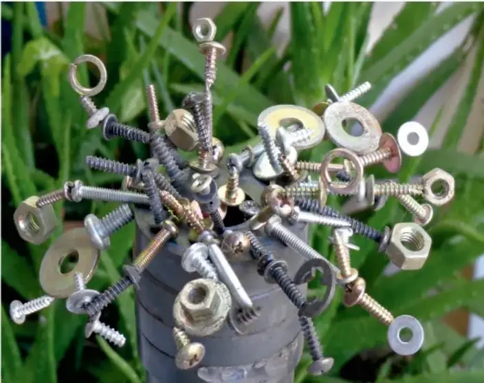 Fotografía de un bote de plástico lleno de tornillos, tuercas, rondanas y clavos que sobresalen, con plantas verdes de fondo.
