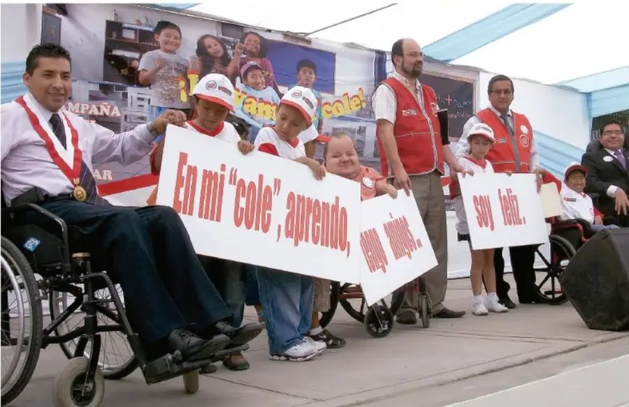 Fotografía de un acto comunitario escolar: niños y adultos, varios en sillas de ruedas, sostienen letreros que dicen «En mi "cole", aprendo...» y «soy feliz». Promueve la convivencia inclusiva.
