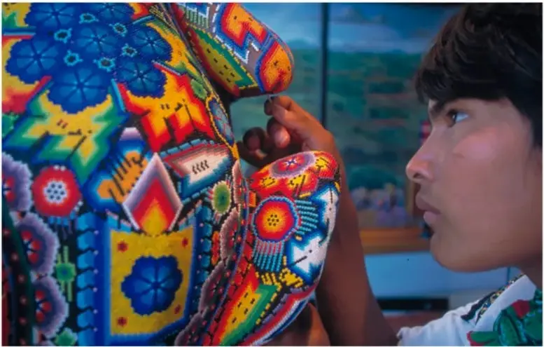 Fotografía de un artesano huichol trabajando una pieza de Chaquira muy colorida (azules, rojos, amarillos, verdes) con motivos florales y geométricos; leyenda: Huichol, Tepic, Nayarit, fotografía de Carlos Sánchez Pereira, Banco de imágenes Conabio.