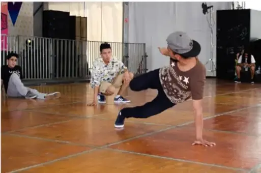 Jóvenes practicando breakdance o danza urbana en un piso de madera, uno de ellos apoyado en una mano en una posición acrobática; ejemplifica la apropiación de una cultura física.