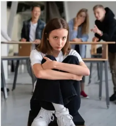 Fotografía de una adolescente sentada en el suelo con los brazos cruzados, con expresión de incomodidad; al fondo, tres compañeros la observan. Representa situaciones de riesgo o exclusión en la escuela.