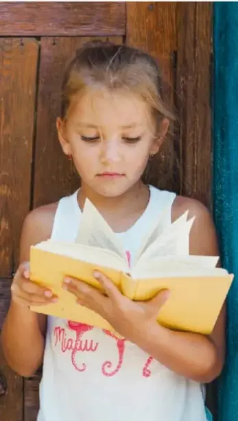 Fotografía de una niña leyendo un libro amarillo junto a una puerta de madera.