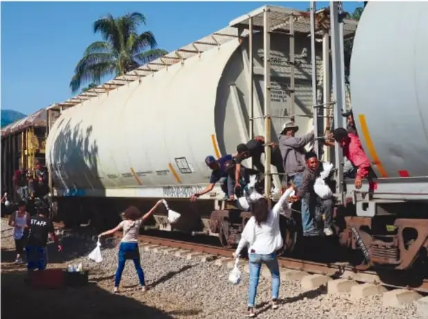 Fotografía de personas subidas a un tren de carga, conocida como 'La Bestia', usada por migrantes para desplazarse; algunas personas en el suelo les lanzan bolsas, posiblemente con alimentos.