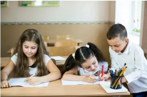 Fotografía de tres estudiantes de primaria escribiendo en sus cuadernos sobre una mesa de madera en el salón de clases.