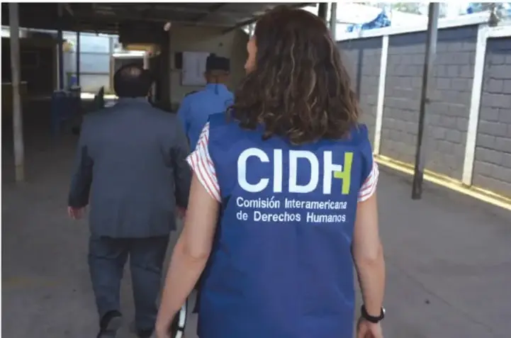 Fotografía de una persona de espaldas con chaleco azul que dice 'CIDH, Comisión Interamericana de Derechos Humanos', caminando por el pasillo de una escuela junto a otras personas.