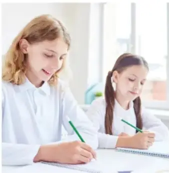 Fotografía de dos niñas de primaria con uniforme blanco, sentadas en un aula escribiendo con lápiz verde en sus cuadernos.