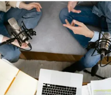 Fotografía cenital de un estudio de grabación: dos personas sentadas frente a micrófonos de estudio con computadora portátil y cuaderno sobre la mesa, ilustrando una entrevista o programa de radio.