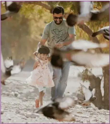 Fotografía de un papá con lentes y camiseta gris que juega con su hija pequeña de vestido blanco entre palomas que vuelan, en un paseo al aire libre.