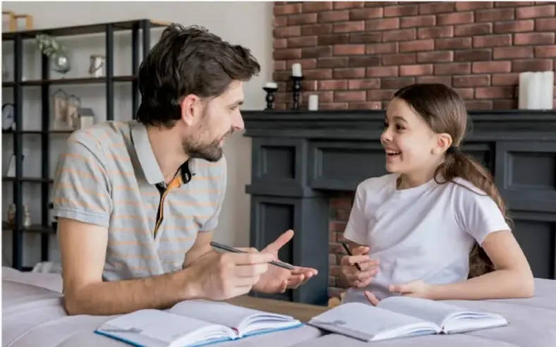 Fotografía de un adulto y una niña sentados frente a una mesa con cuadernos abiertos, conversando y sonriendo. Al fondo hay una estantería y una chimenea de ladrillo.