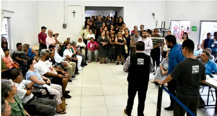 Fotografía de una asamblea comunitaria: un grupo numeroso de personas sentadas en círculo en un salón, atentas a dos jóvenes de pie que conversan, ilustrando una actividad comunitaria planificada.