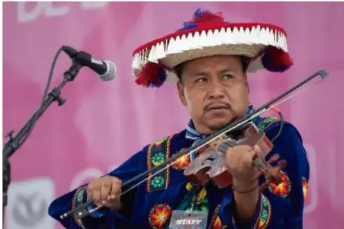 Fotografía de un músico indígena con sombrero tradicional bordado y traje típico, tocando el violín frente a un micrófono, en un escenario con fondo rosa.