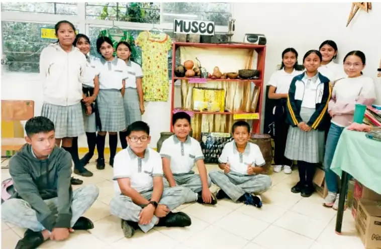 Fotografía de un grupo de estudiantes de secundaria con uniforme, dentro de un salón-museo escolar, junto a estantes con artesanías, jarrones y un letrero que dice "MUSEO".