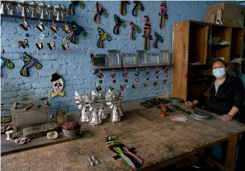 Taller artesanal con pared de ladrillo azul, adornada con figuras decorativas colgantes. Una persona con cubrebocas trabaja en una mesa de madera con herramientas y piezas metálicas. Se observan productos terminados y cajas al fondo.