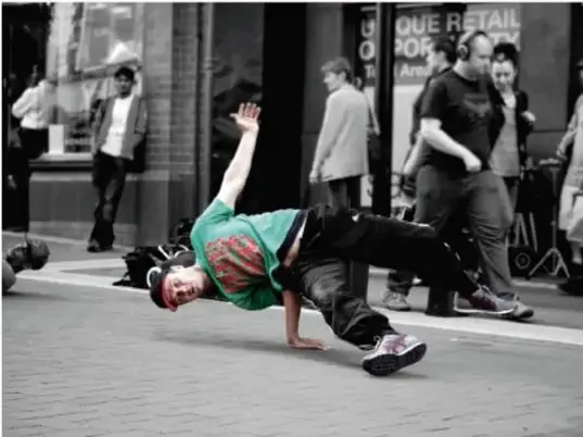 Fotografía en blanco y negro de un joven bailando breakdance en una calle urbana, con transeúntes al fondo.