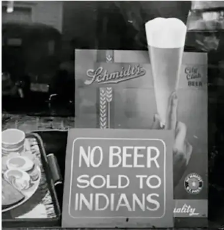Fotografía documental en blanco y negro de un establecimiento con un cartel que dice "NO BEER SOLD TO INDIANS" (no se vende cerveza a indios), ejemplo de prácticas racistas discriminatorias.
