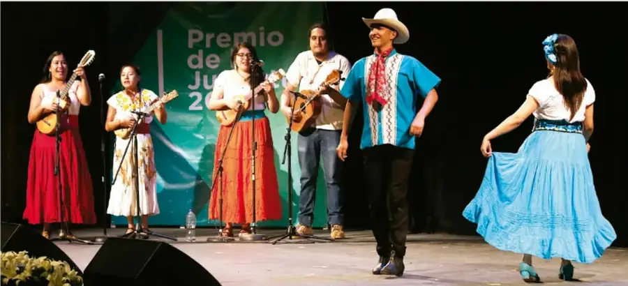 Fotografía de un grupo de jóvenes en un escenario: tres mujeres con vestidos largos de colores tocando ukuleles, un hombre con guitarra, un hombre con sombrero y camisa azul al centro y una mujer con falda azul bailando; detrás, un telón con el texto 'Premio de Jóvenes'.