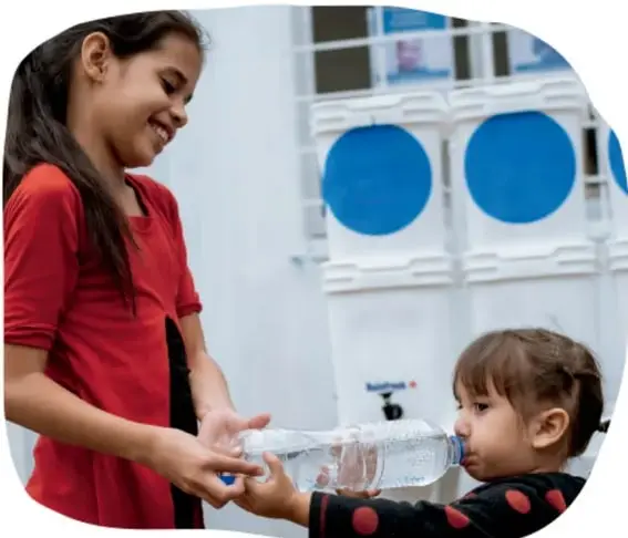 Fotografía de una niña en rojo dando agua de una botella a una niña menor, ilustrando la acción adecuada al contexto.