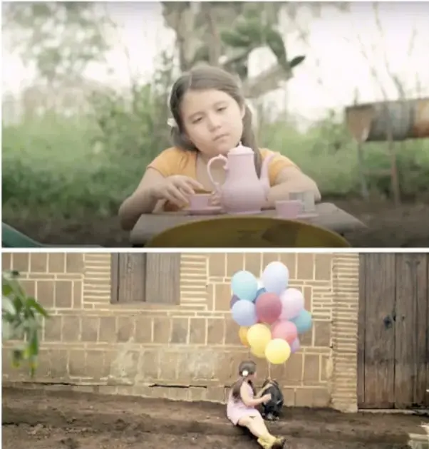 Fotograma de Valentina sentada junto a un cerdo con un ramo de globos de colores frente a una pared de block.