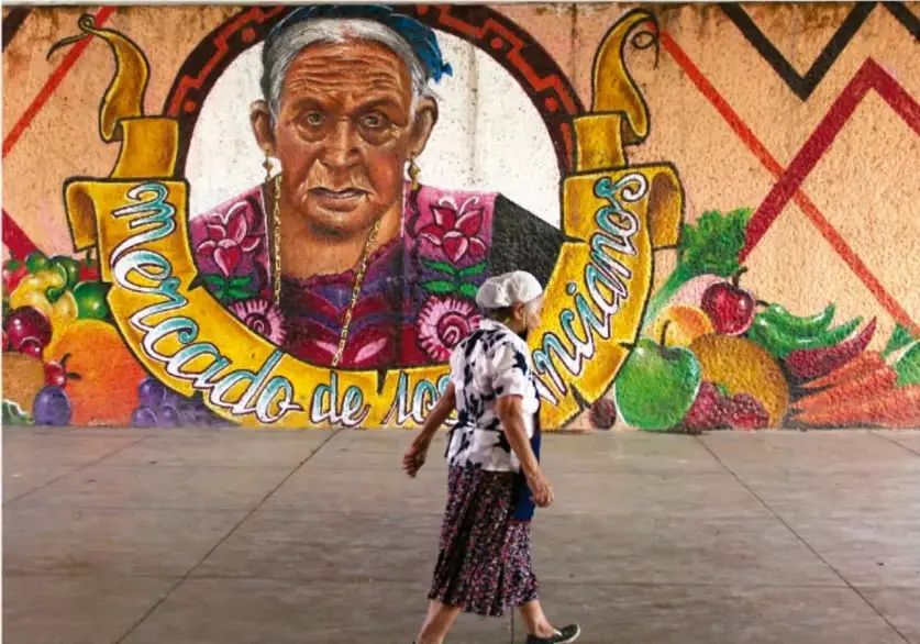 Mural comunitario titulado "Mercado de los mariscos" con el retrato de una mujer mayor, frutas, verduras y motivos geométricos; una persona camina frente al mural.